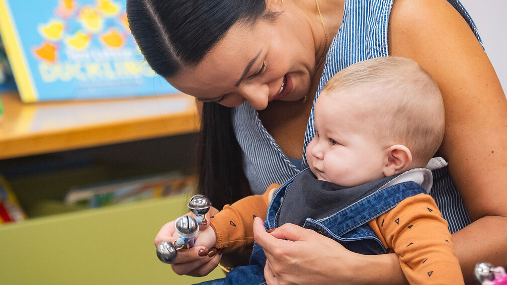A woman smiling while holding a baby in her lap and using a rattle during a story time session