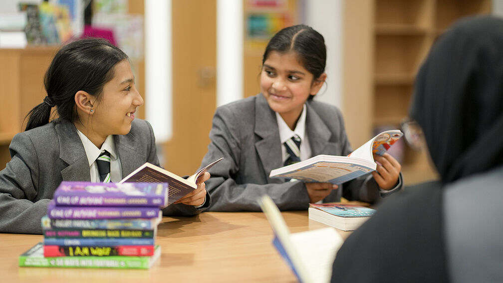 A photo of two girls in school uniform sitting at a table with books on it, each holding a book, looking at each other and smiling