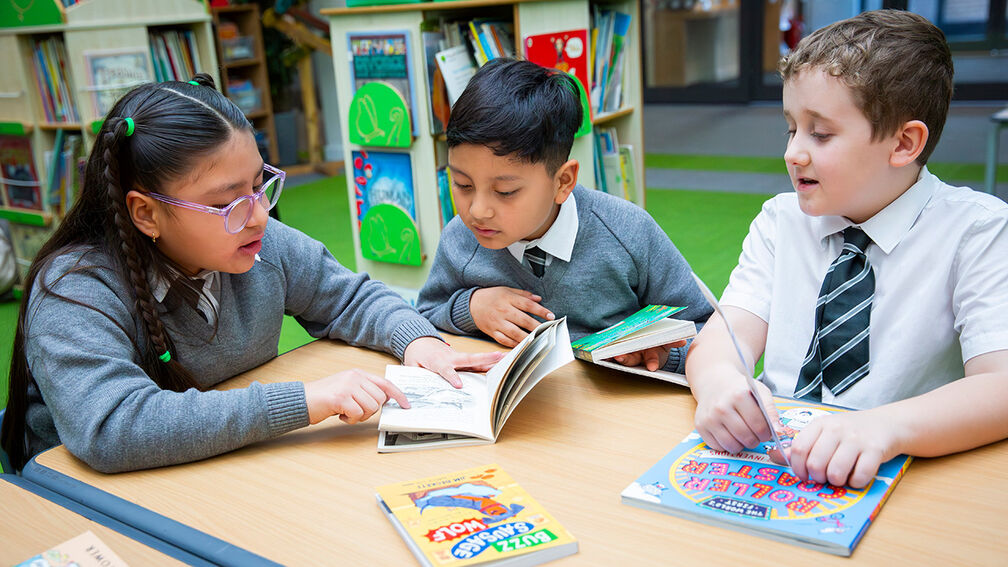 A photo of three primary aged children - a girl and two boys - wearing school uniform, sitting at a desk and looking at books together