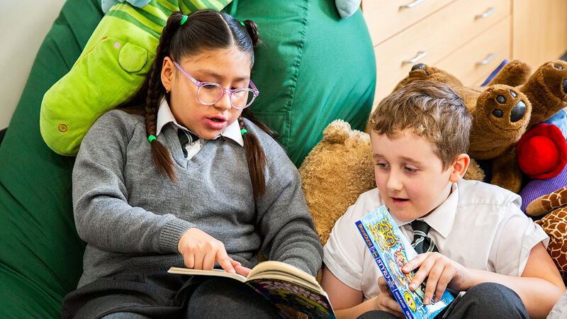 A primary aged girl and boy in school uniform - the girl is pointing something out on a page of a book to the boy, who is holding a book in his hands
