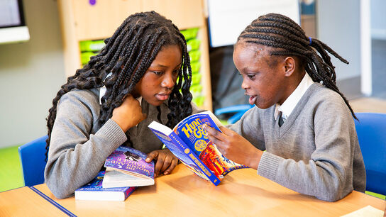Two primary aged girls in school uniform sitting at a table and looking at books together