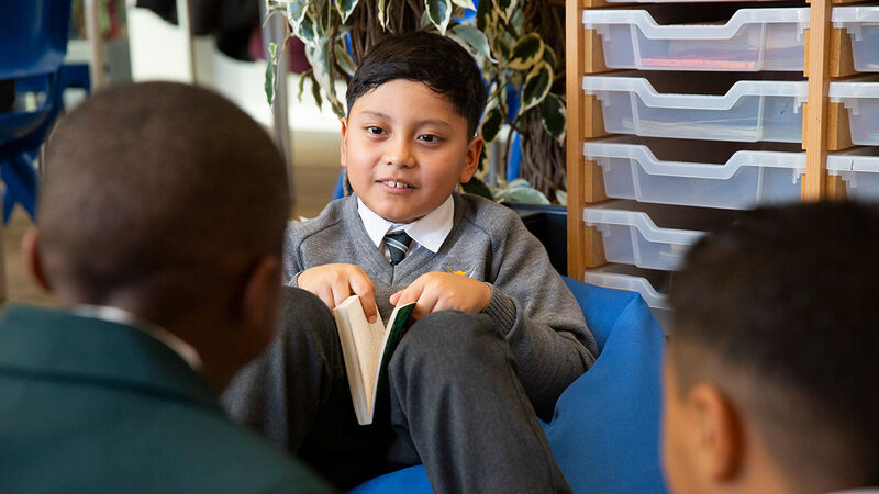 A primary aged child sitting in a beanbag and holding a book, smiling at his friends