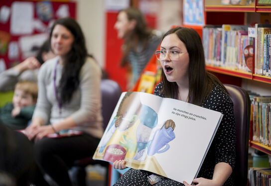A librarian reading out a picture book to a group of children with an animated expression on her face.