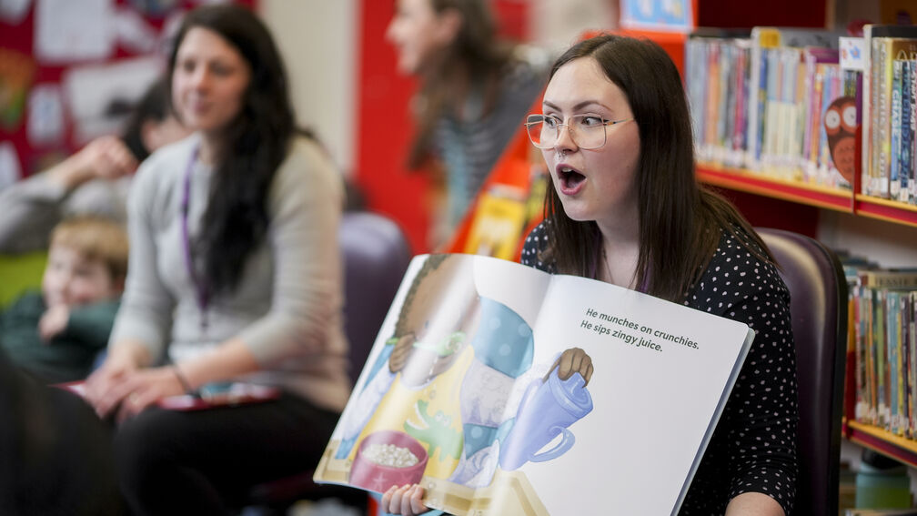 A librarian reading out a picture book to a group of children with an animated expression on her face.