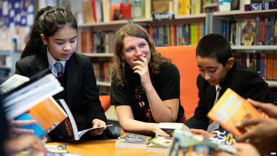 A smiling school librarian sitting in a library with a group of students, who are sitting around her all looking at books