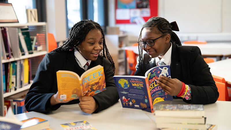Two girls smiling while looking at books together, sitting at a desk in their school library with piles of books in front of them