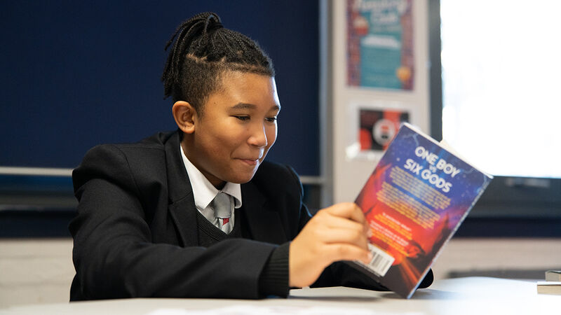 A boy smiling while reading at a desk in a school classroom