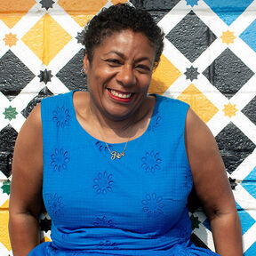 Waterstones Children's Laureate Patrice Lawrence smiling as she sits in front of a wall painted in colourful diamonds