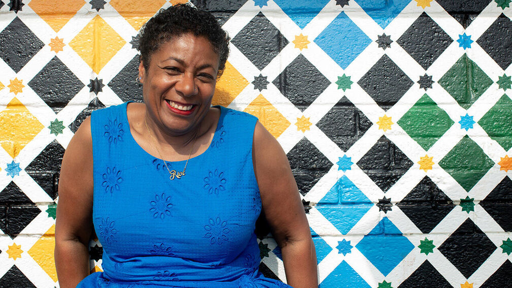 Waterstones Children's Laureate Patrice Lawrence smiling as she sits in front of a wall painted in colourful diamonds