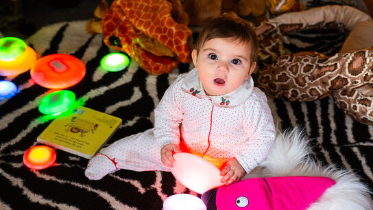 A baby sitting on the floor at a storytelling session in Knowsley, surrounded by lights, toys and rugs - the baby is very cute and looking at the camera with its mouth open