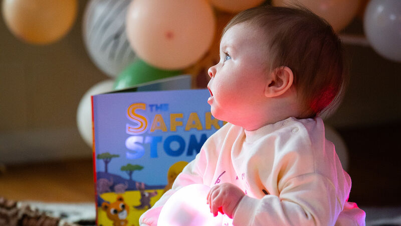 A baby sitting on the floor at a storytelling session, looking up with an open mouth. The book The Safari Stomp is next to them and balloons can be seen in the background.