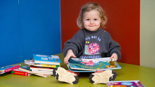 A photo of a young child in Barnsley, smiling at the camera as she sits on the floor with books on her lap and piled up next to her