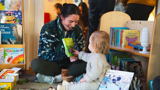 A woman and a baby sitting on the floor in a library or children's centre, surrounded by bookshelves and book cases. The woman is looking excited as she shows a book to the baby, who points at its front cover