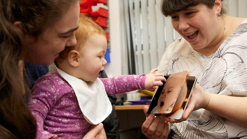 A baby in a purple top reaches for a picture book, captivated by a photograph of a yawning baby. A smiling woman holds the book, while another caregiver watches warmly, sharing the moment.