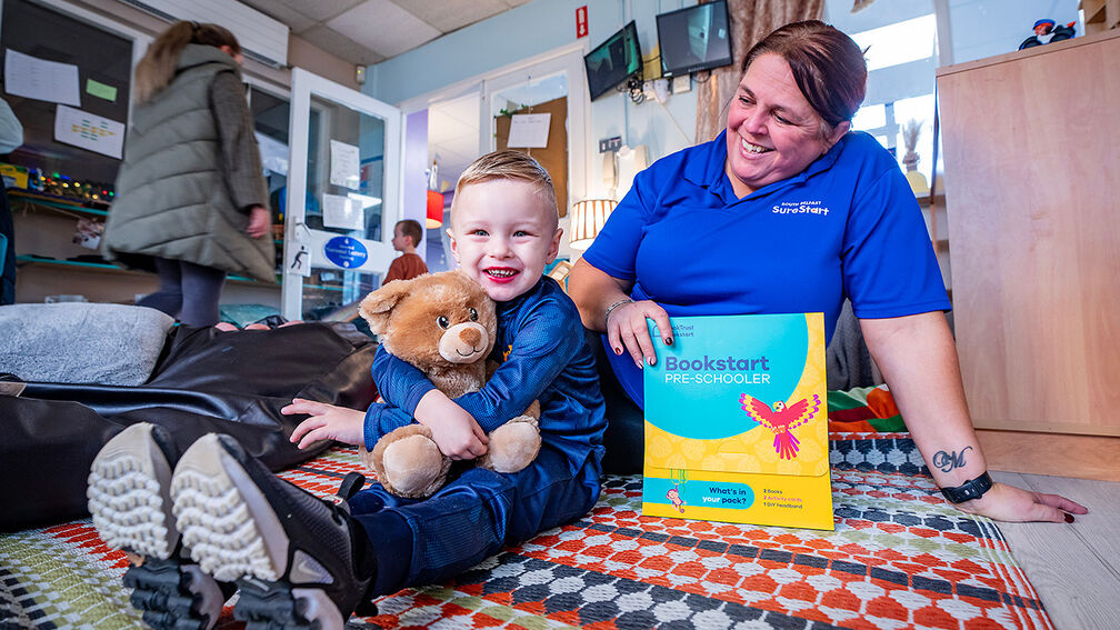 A child hugging a Build-a-Bear bear at South Belfast Sure Start as a woman smiles and holds a Bookstart pack