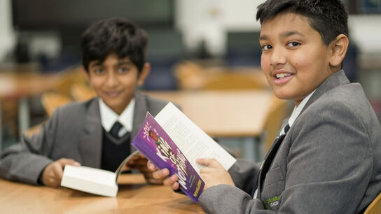Two boys in a classroom setting are smiling at the camera while reading books