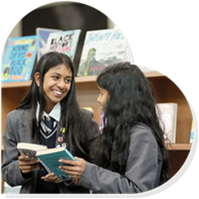 Two secondary school age girls are smiling at each other while holding books in front of some bookshelves