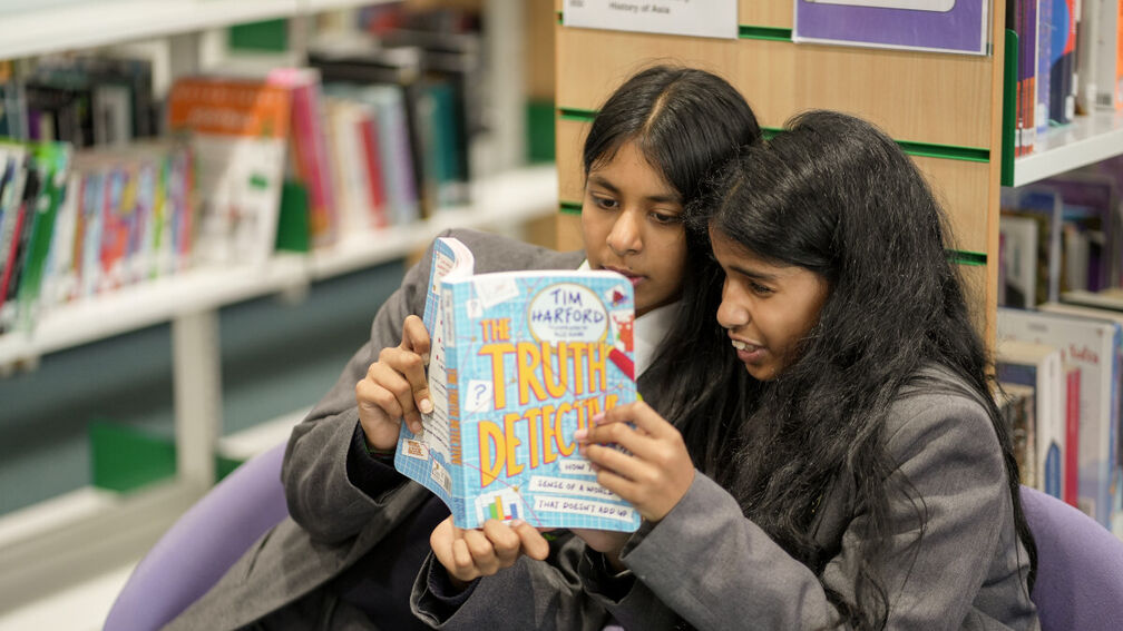 Two 11-year-old girls are sitting together sharing a book in a school library setting