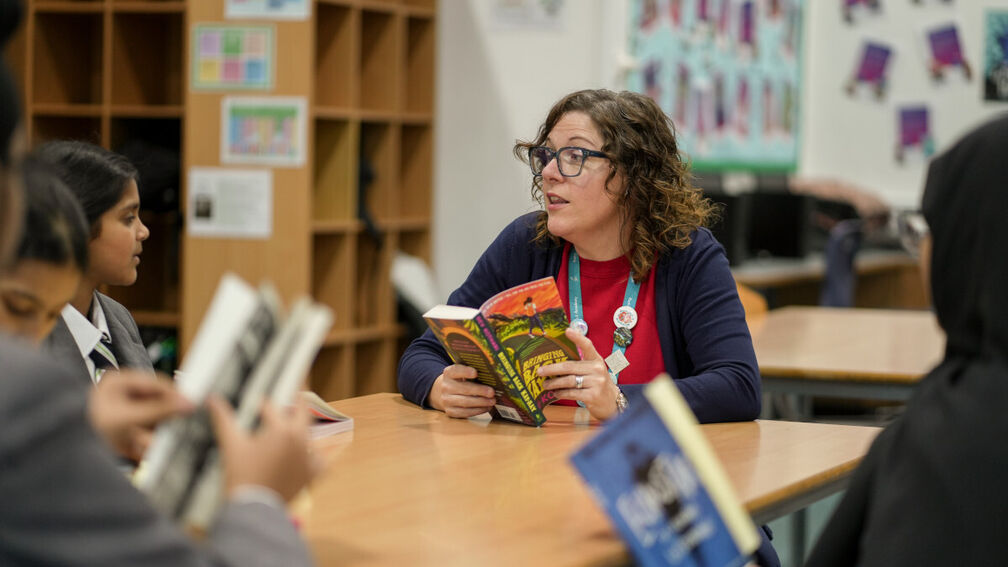 A teacher is sitting at the head of a table of 12 year old school children in a school library setting. They are all reading their own copies of a book.