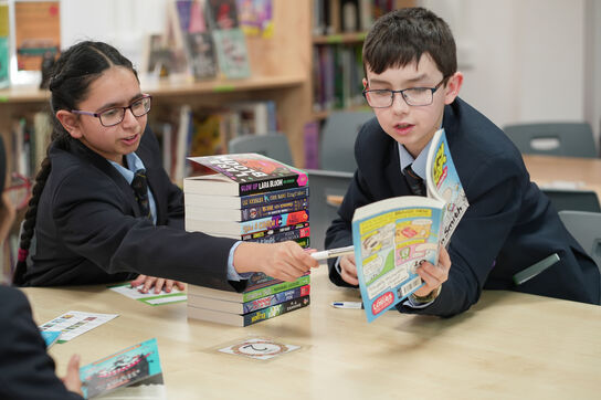 A primary school aged boy is in a school library showing a classmate something in a Bookbuzz book.