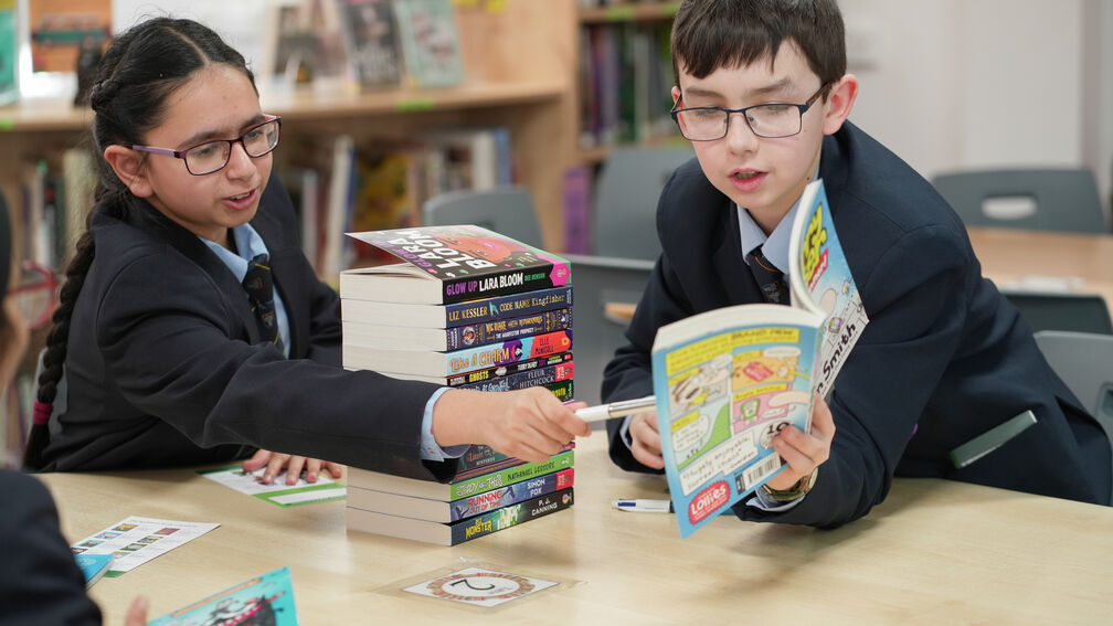 A primary school aged boy is in a school library showing a classmate something in a Bookbuzz book.