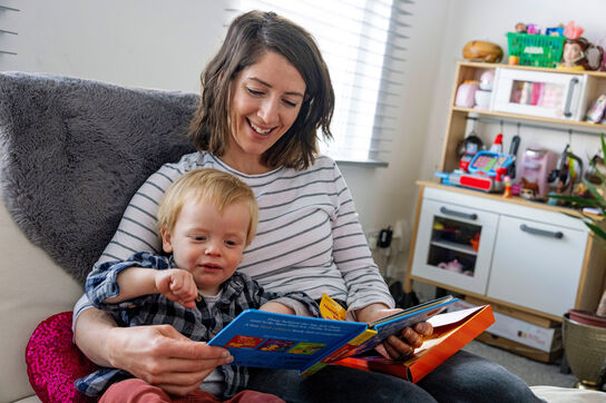 A mother and her young child share a joyful moment reading together on a cosy sofa. The toddler, reaches out excitedly towards the colourful board book, fully engaged in the story.
