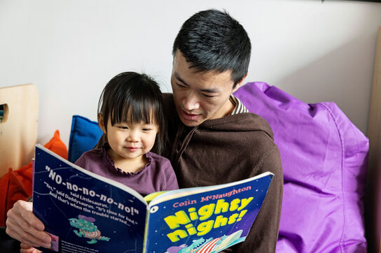 A father and his young daughter sit together in a cosy reading nook, surrounded by colourful cushions reading the book 'Nighty Night' by Colin McNaughton