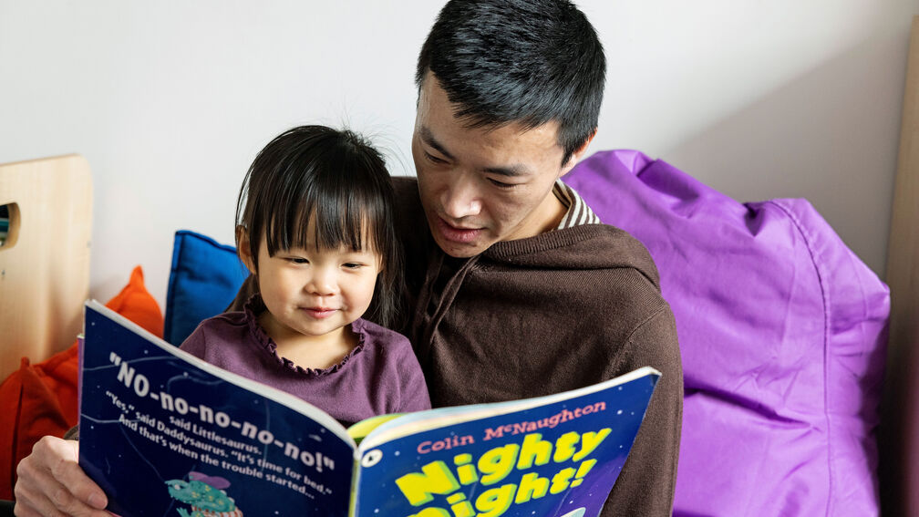 A father and his young daughter sit together in a cosy reading nook, surrounded by colourful cushions reading the book 'Nighty Night' by Colin McNaughton