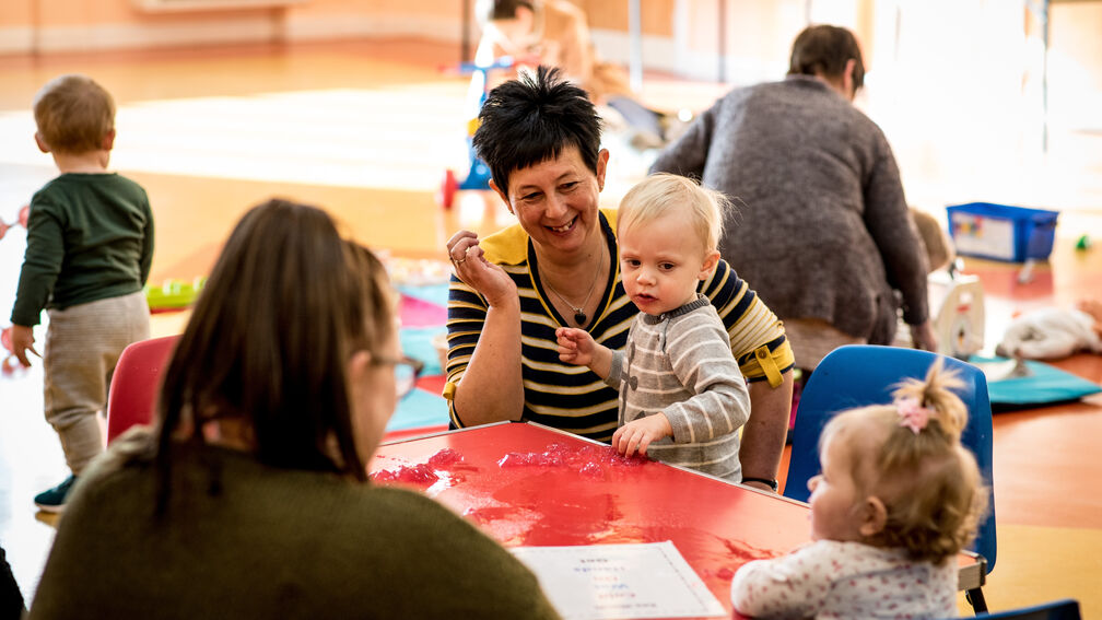A toddler is with his carer in a childcare setting, with other carers and toddlers around