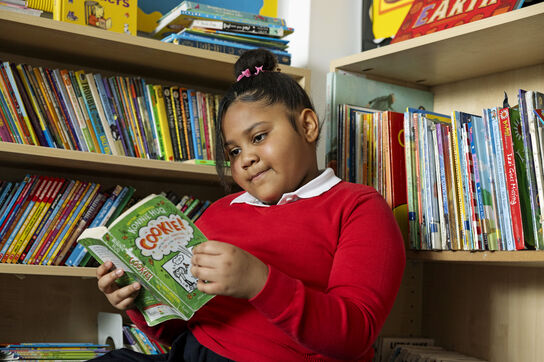A primary school aged girl is sitting reading a book surrounded by bookshelves and books