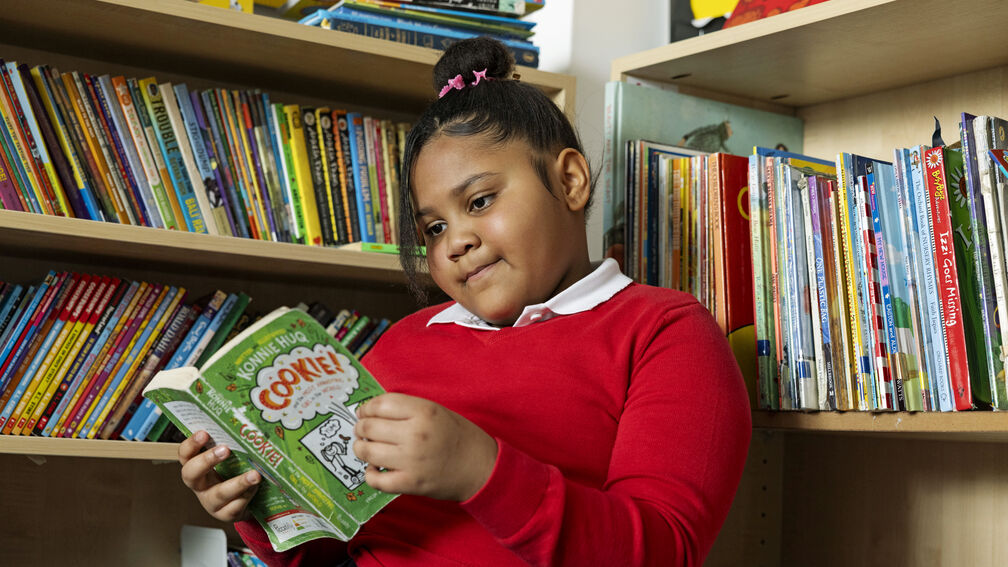 A primary school aged girl is sitting reading a book surrounded by bookshelves and books
