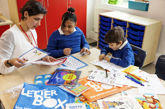 Two primary school aged children, a girl and a boy, are sitting with a teacher at a table surrounded by the contents of their blue Letterbox Club parcel