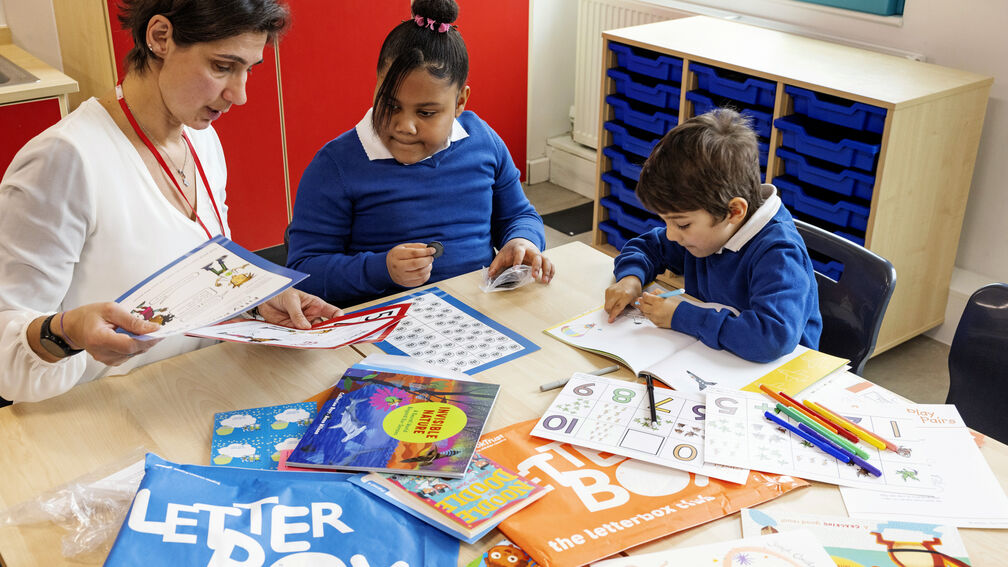 Two primary school aged children, a girl and a boy, are sitting with a teacher at a table surrounded by the contents of their blue Letterbox Club parcel