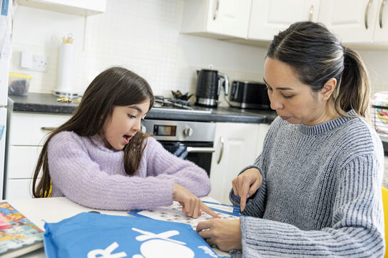 A primary school aged girl is sitting in a kitchen with her female carer and doing activities from the blue Letterbox Club parcel