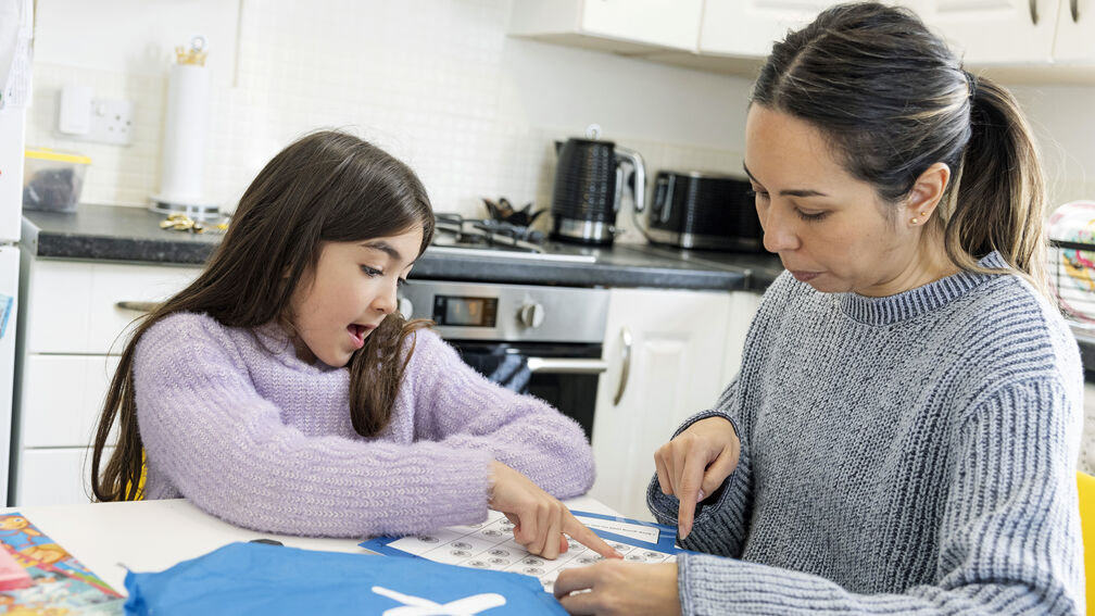 A primary school aged girl is sitting in a kitchen with her female carer and doing activities from the blue Letterbox Club parcel