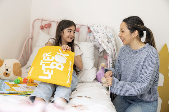 A primary school aged girl is sat on her bed opening her yellow Letterbox Club parcel and smiling, while her female carer watches.