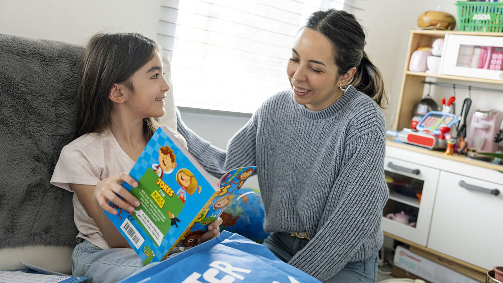 A smiling girl is reading the contents of her Letterbox Club pack with  her female carer sitting next to her
