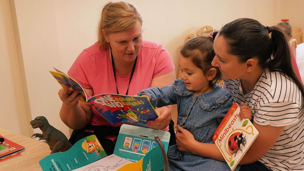two people and a child sitting down on the floor with the child reading and pointing at an open book