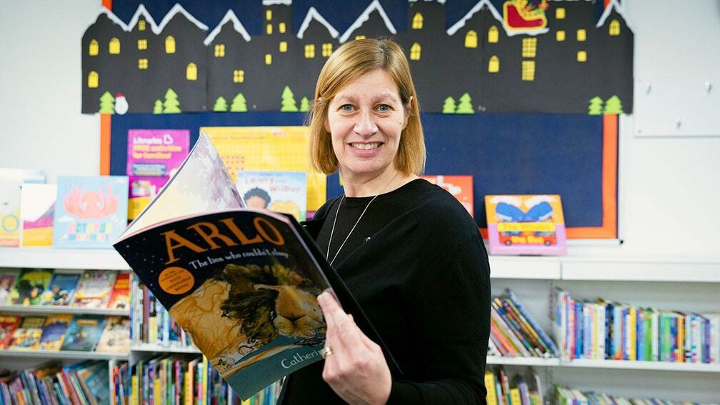 Donna in the library holding a book, looking to camera and smiling.