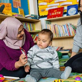 A mother shows a baby a finger puppet with a practitioner overseeing the interaction.