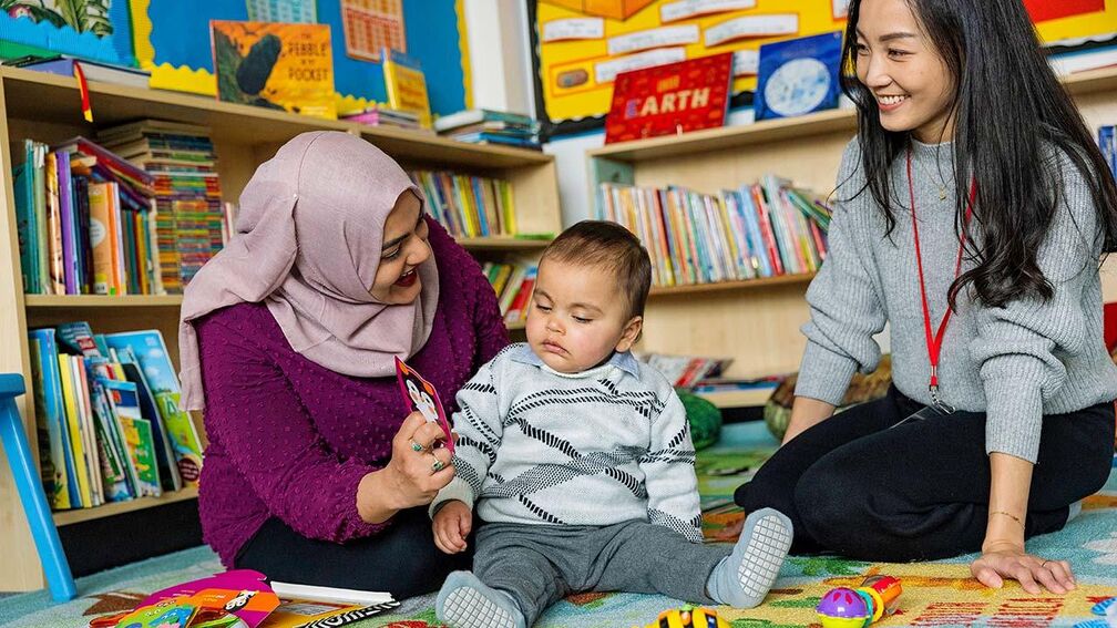 A mother shows a baby a finger puppet with a practitioner overseeing the interaction.