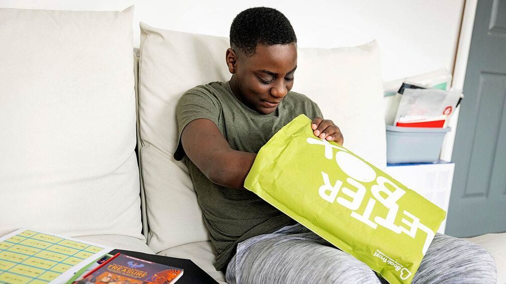 A 10 year old boy is sitting on a sofa and opening a green Letterbox Club parcel and smiling