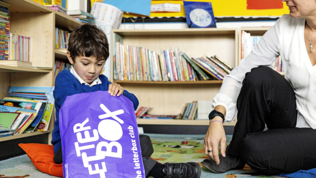 A primary school aged boy is opening his purple Letterbox Club parcel while his female teacher watches him.