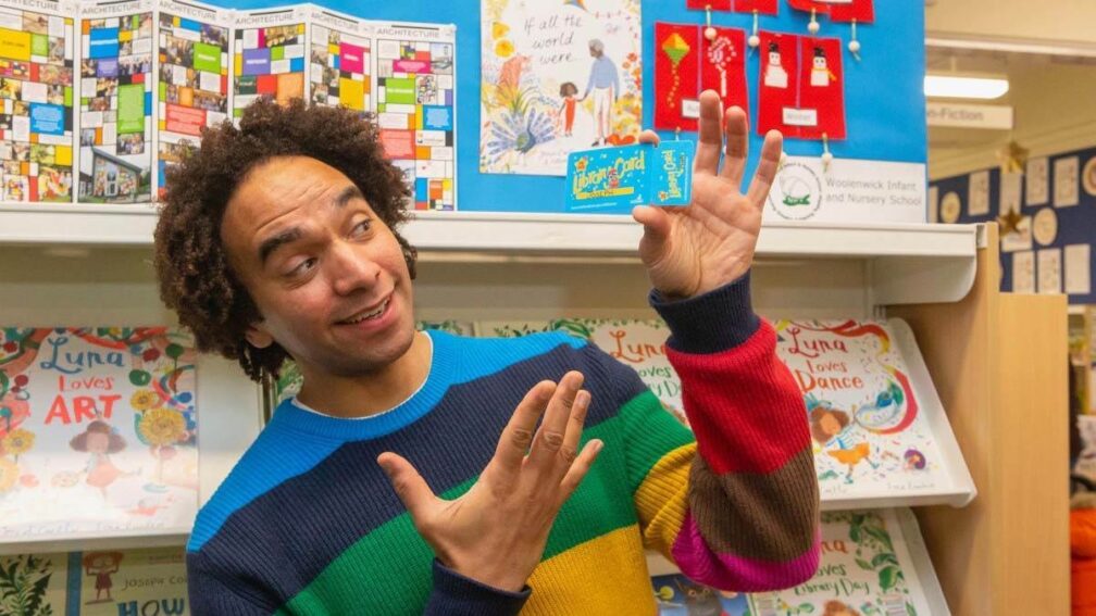 Joseph Coelho is standing in a library in a brightly coloured jumper, in front of a rack of books, and holding up a library card