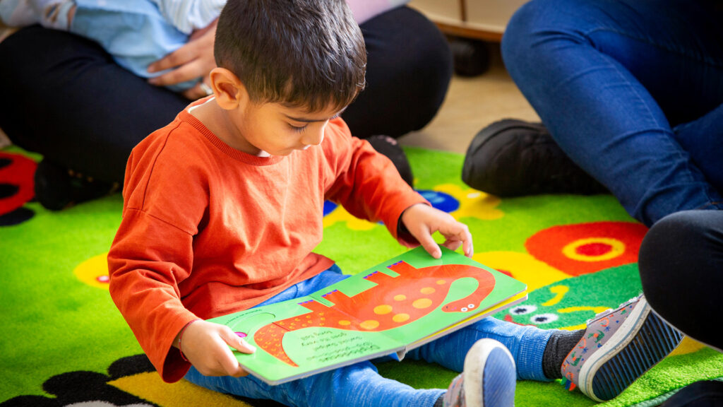 A child sitting on the floor looking at a book with an illustration of a dinosaur.