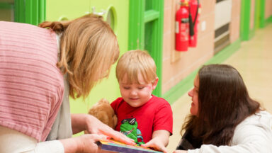 Two early-years practitioners are showing a young boy a book in a nursery setting