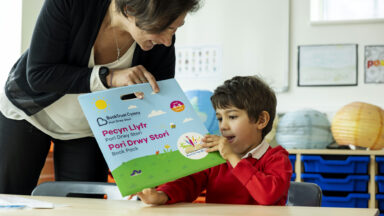 A young primary school aged boy is being given a Pori Drwy Story book pack by his teacher, they are both smiling.
